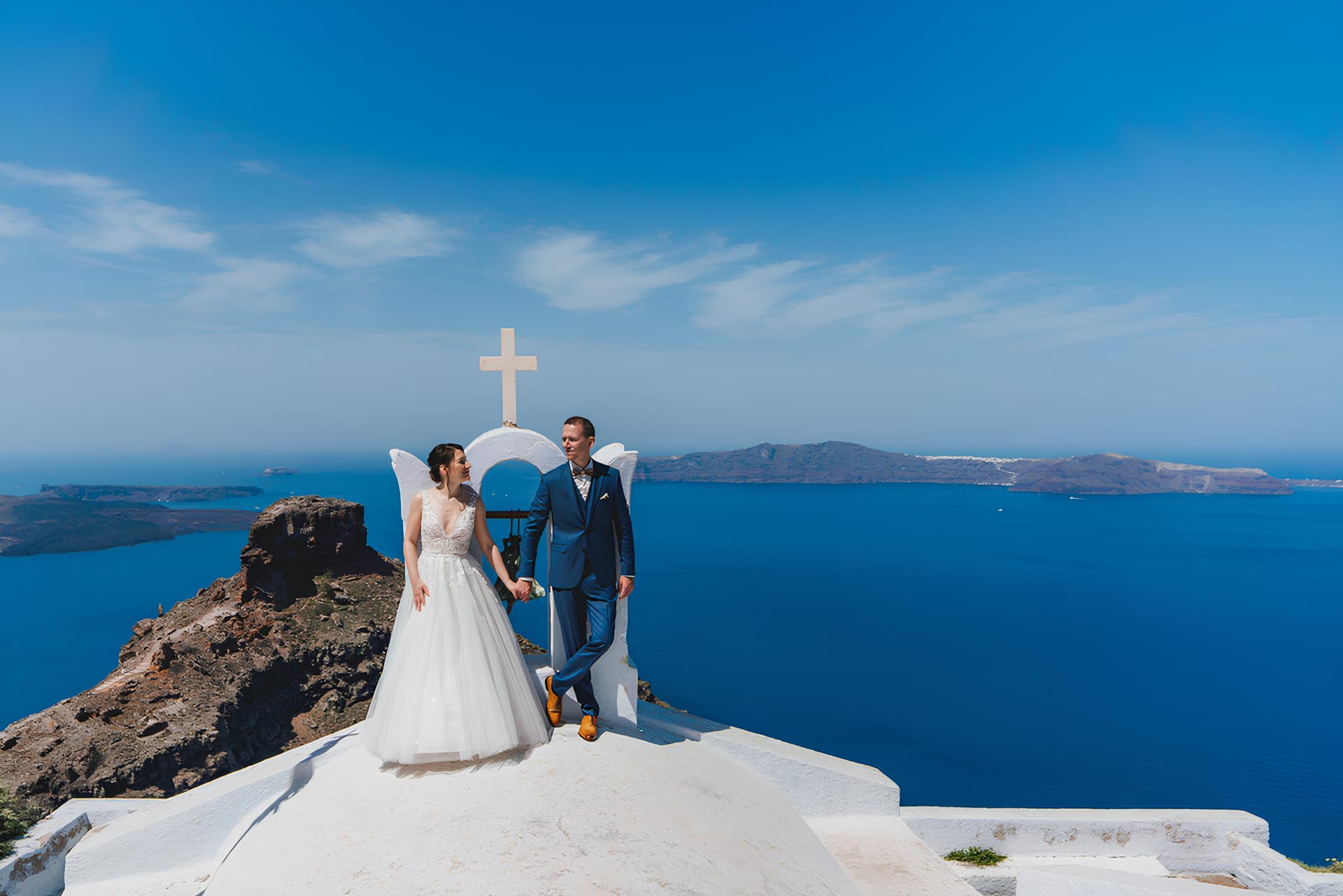 A bride and groom holding hands on the Santorini caldera, the Aegean sea behind them